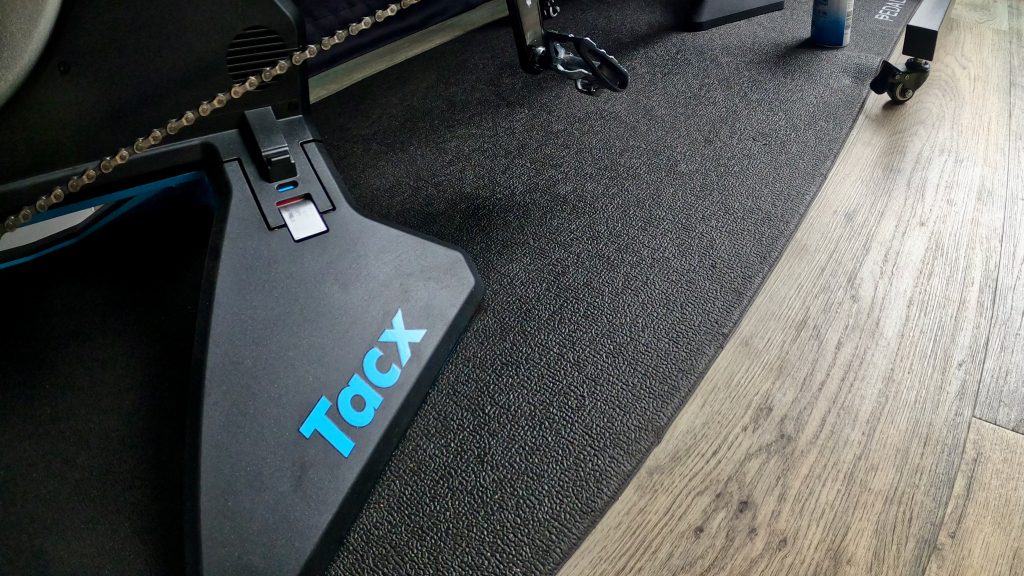Close-up of a Tacx smart trainer's black frame with the "Tacx" logo in blue, bike chain and pedal visible above, set on a black training mat over wood flooring.