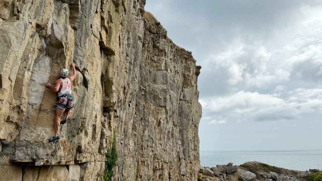 Climber in a white helmet reaching for a hold on the limestone wall of I Thought You Had It at Winspit, with the sea behind